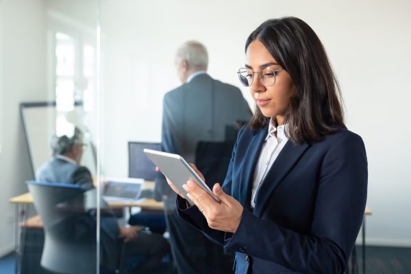 Focused office lady in glasses using tablet while two mature businessmen discussing work behind glass wall. Copy space. Communication concept